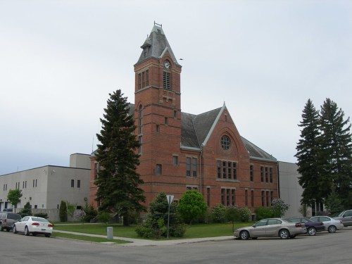 Historic brick building with a clock tower, surrounded by trees and modern structures, under a cloudy sky.