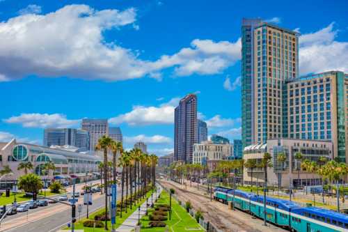 A vibrant cityscape featuring modern buildings, palm trees, and a train line under a bright blue sky with clouds.