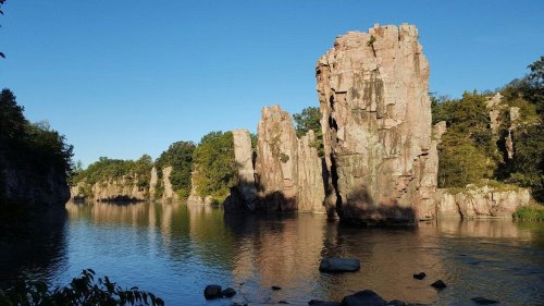 Rock formations rise above a calm river, surrounded by lush greenery under a clear blue sky.