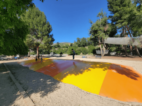 A colorful inflatable bounce pad in a sandy area, with children playing and trees in the background under a clear blue sky.