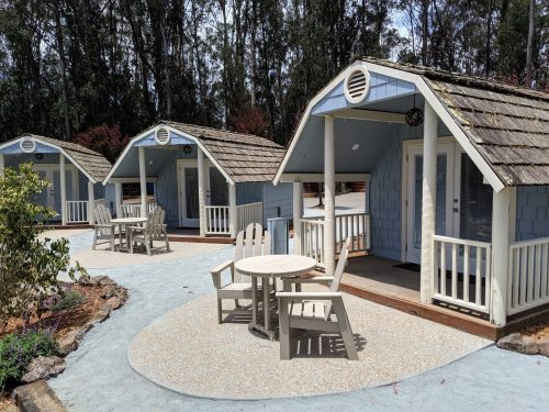 Three small blue cabins with wooden roofs, surrounded by trees, and a circular seating area with tables and chairs.