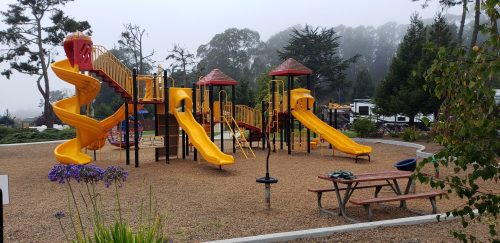 A colorful playground with yellow slides and climbing structures, surrounded by trees and a foggy atmosphere.