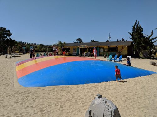 A colorful inflatable bounce pad on a sandy beach, with children playing and a building in the background.