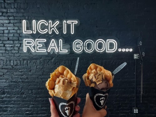 Two hands hold waffle cones filled with ice cream, against a black wall with the neon text "LICK IT REAL GOOD."