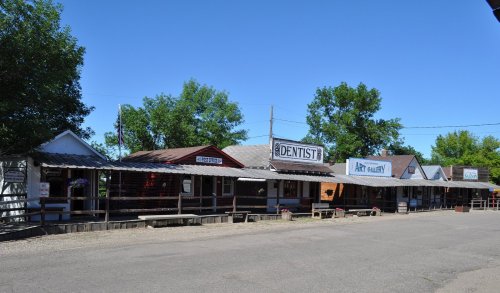 A row of quaint storefronts, including a dentist and art gallery, under a clear blue sky with trees in the background.