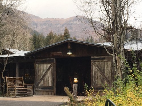 A rustic wooden building with large doors, surrounded by trees and mountains in the background, on a cloudy day.