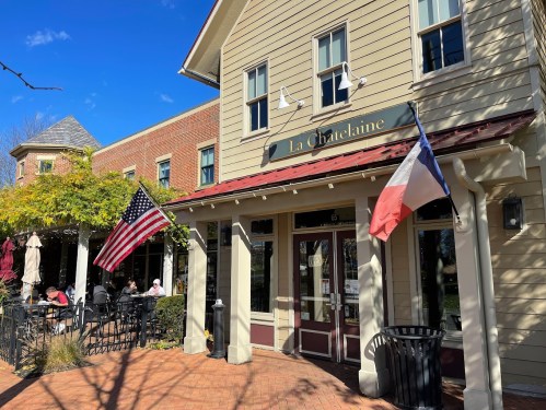 A charming café named "La Châtelaine" with outdoor seating, featuring American and French flags, under a clear blue sky.