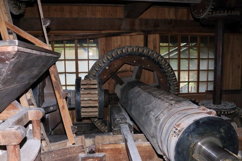 Interior of a vintage mill showing large gears and machinery, with wooden structures and windows in the background.
