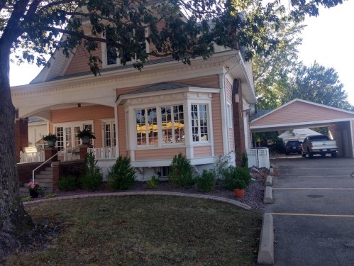 A charming two-story house with a porch, surrounded by greenery and a driveway leading to a carport.
