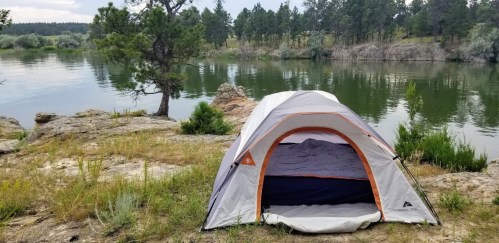 A tent set up by a calm river, surrounded by greenery and trees, with rocky terrain in the background.