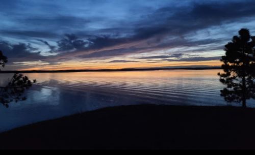 A serene lake at sunset, with colorful clouds reflecting on the water and silhouettes of trees in the foreground.