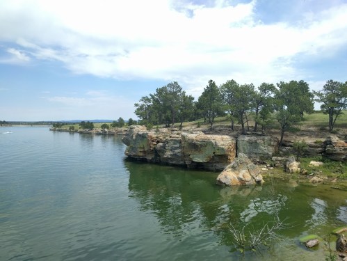 A serene lake scene with rocky cliffs and green trees under a partly cloudy sky.