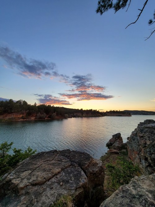 A serene river scene at sunset, with colorful clouds reflecting on the water and rocky shores in the foreground.