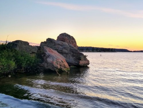 A rocky shoreline at sunset, with calm water reflecting the colorful sky and distant trees on the horizon.