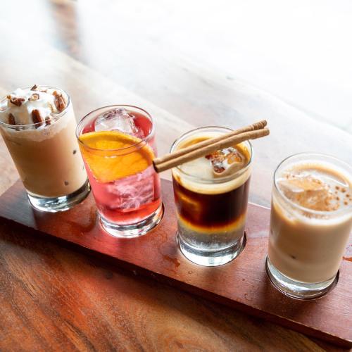A wooden tray holds four colorful drinks: a creamy iced coffee, a fruity cocktail, a dark espresso, and a milky beverage.