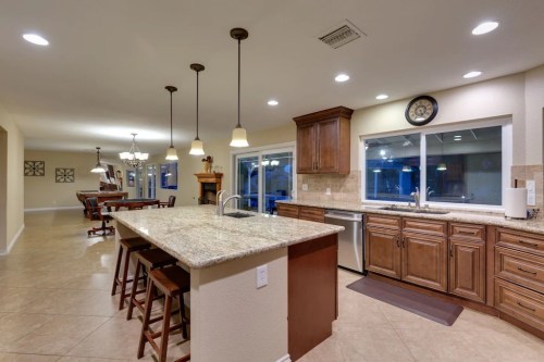 Modern kitchen with granite island, wooden cabinets, and large windows, featuring bar stools and warm lighting.