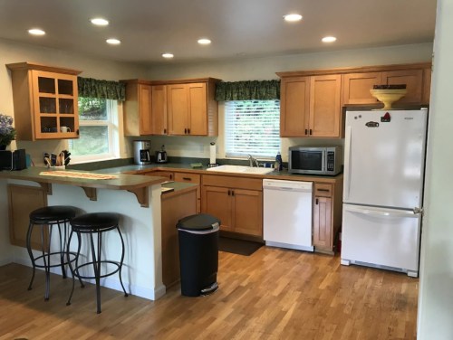 Bright kitchen with wooden cabinets, white appliances, and a bar with stools, featuring a window and wooden flooring.