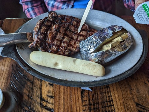 A grilled steak on a plate with a baked potato wrapped in foil and a breadstick on the side.