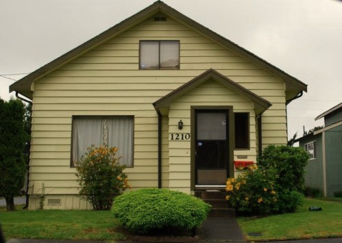 A light green house with a front porch, numbered 1210, surrounded by shrubs and flowers on a cloudy day.