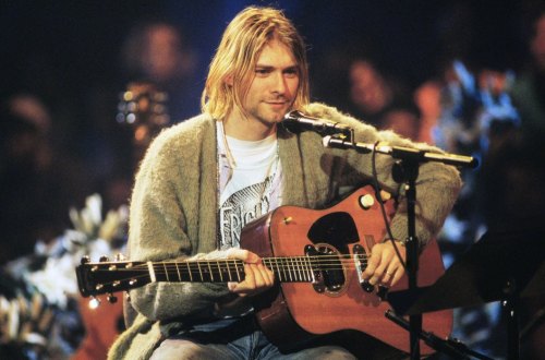 Kurt Cobain sitting with an acoustic guitar, wearing a cozy sweater, performing on stage.