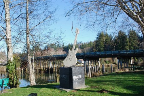 A guitar sculpture stands on a pedestal by a river, with a bridge and trees in the background under a clear blue sky.