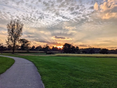 A serene landscape at sunset, featuring a path, grassy fields, and trees under a colorful sky with clouds.