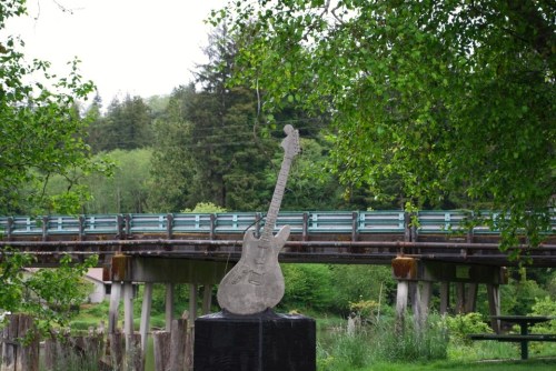 A guitar sculpture stands in front of a wooden bridge surrounded by lush greenery.