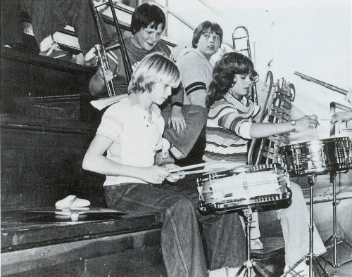 Four young musicians play instruments in a school setting, with a drummer, a violinist, and others seated nearby.