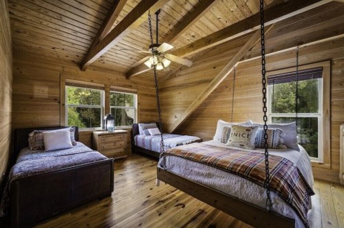 Cozy wooden bedroom with two hanging beds, a nightstand, and large windows overlooking greenery.