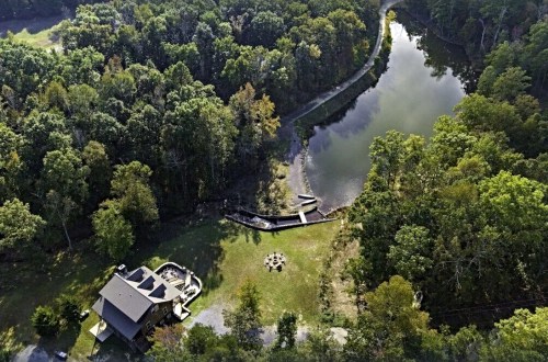 Aerial view of a house by a pond, surrounded by trees, with a fire pit and a small dock visible.