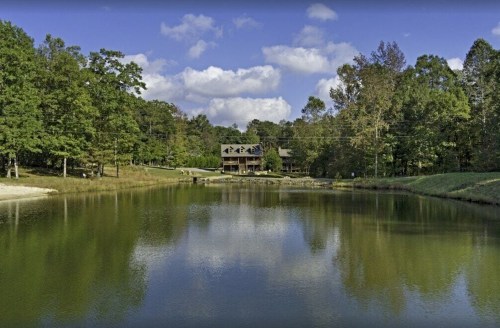 A serene lake surrounded by trees, with a house visible on the shore under a partly cloudy sky.