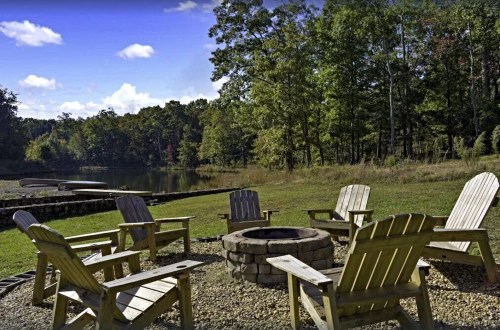 A serene lakeside scene featuring wooden chairs arranged around a stone fire pit, surrounded by trees and blue skies.
