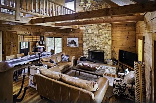 Cozy wooden cabin interior featuring a stone fireplace, leather sofas, and a rustic dining area with natural light.