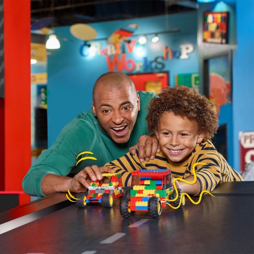 A joyful man and boy play with colorful toy cars made of building blocks in a vibrant workshop setting.