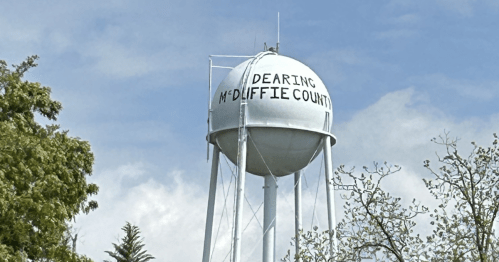 Water tower with "Dearing McDuffie County" painted on it, surrounded by trees and a blue sky with clouds.