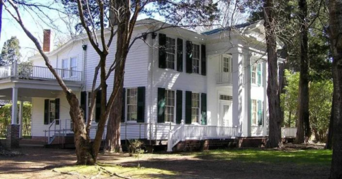 A large white house with black shutters, surrounded by trees and greenery, featuring a porch and multiple windows.