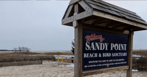 Sign for Sandy Point Beach & Bird Sanctuary, with a sandy area and marshland in the background under a cloudy sky.