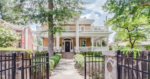 A large, two-story house with a porch, surrounded by greenery and a black iron gate.