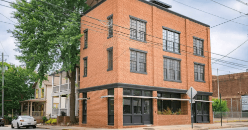 A modern brick building with large windows, situated at a street corner, surrounded by trees and other structures.