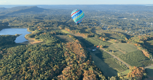 A colorful hot air balloon floats over a scenic landscape of forests, fields, and a lake during autumn.