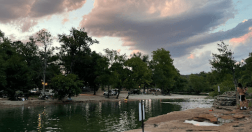 A serene lakeside scene with trees, clouds, and a rocky shore, featuring campers in the background.