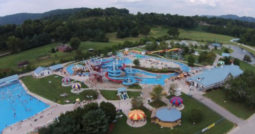 Aerial view of a water park with slides, pools, and colorful umbrellas, surrounded by green hills and trees.