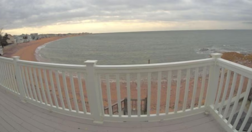 View of a beach with a cloudy sky, seen from a wooden deck with a white railing.