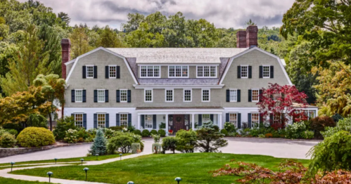 A large, elegant house with a gray exterior, surrounded by lush greenery and colorful trees under a cloudy sky.