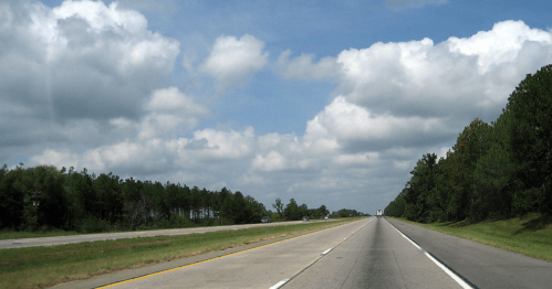 A wide highway stretches into the distance, flanked by green trees and a blue sky with fluffy clouds.