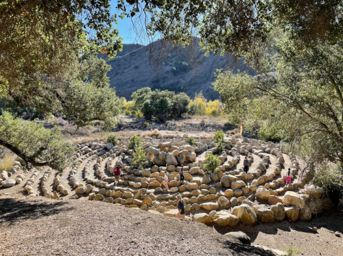 A stone labyrinth surrounded by trees and mountains, with people walking along the paths in a sunny outdoor setting.