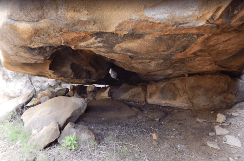 A rocky cave with a large overhang, surrounded by boulders and sparse vegetation.