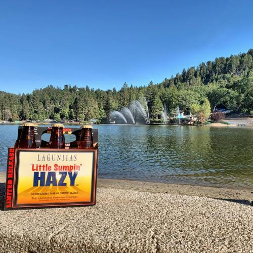 A six-pack of Lagunitas Little Sumpin' Hazy beer sits by a serene lake with fountains and trees in the background.