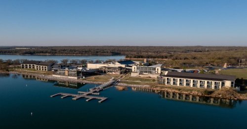 Aerial view of a lakeside resort with modern buildings and a dock, surrounded by trees and water.
