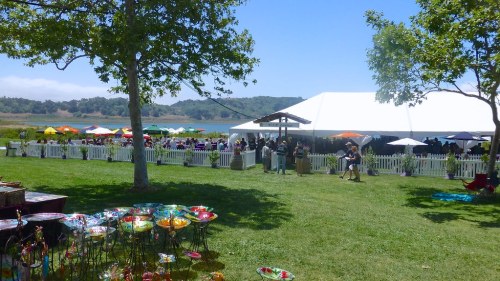 A sunny outdoor event with a large white tent, colorful umbrellas, and people enjoying the scenery by a lake.